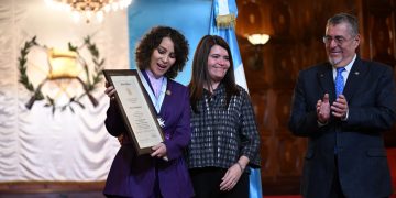 La cantautora Gaby Moreno contempla el reconocimiento que recibe de manos del presidente Bernardo Arévalo y de la secretaria privada de la Presidencia, Glenda Tager. /Foto: Byron de la Cruz