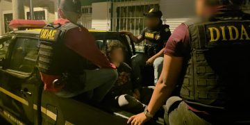 Agentes con chalecos de la DIDAE custodian a un hombre con el rostro difuminado en la parte trasera de una patrulla.