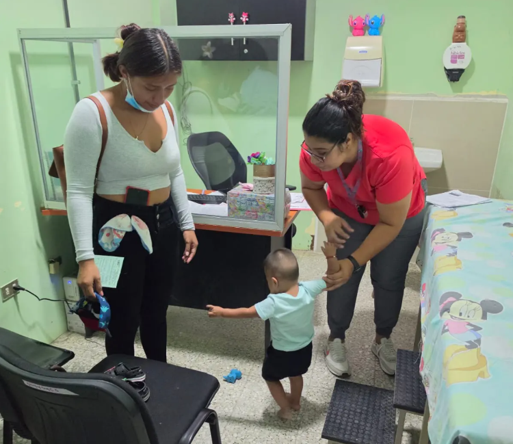 Niño de pie sostiene la mano de una trabajadora de salud mientras una mujer lo acompaña en el consultorio.