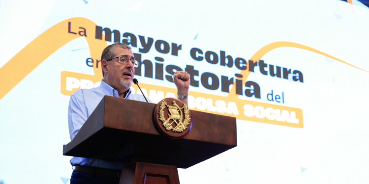 Presidente Bernardo Arévalo habla desde un podio con el escudo nacional, frente a una pantalla sobre la cobertura de Bolsa Social.