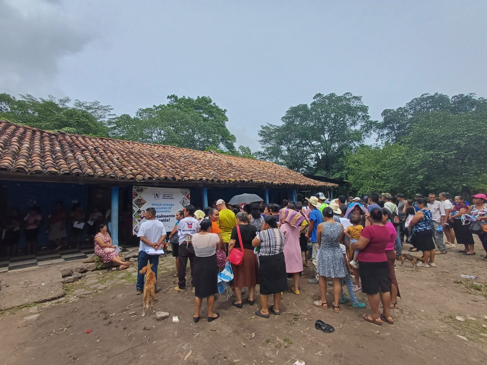 Familias se reúnen frente a un inmueble con techo de tejas y una manta del MAGA que anuncia la entrega gratuita de alimentos.