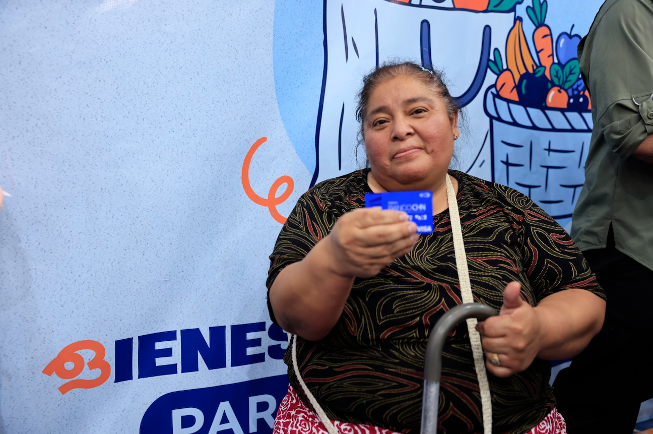 Una mujer muestra una tarjeta azul y levanta el pulgar frente a una manta de Bolsa Social.