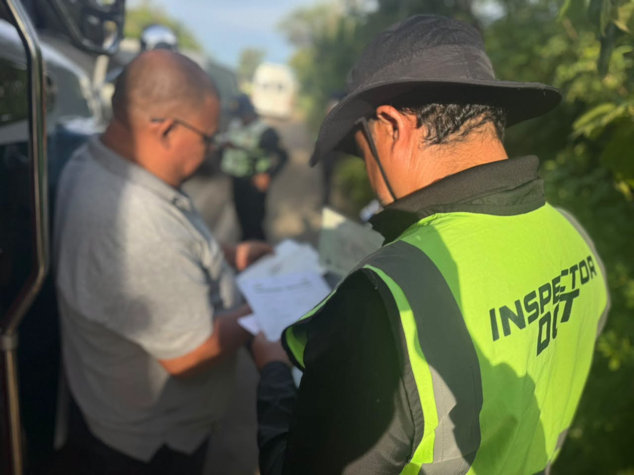 Un inspector de la DGT con chaleco reflectante revisa documentos junto a un hombre al lado de un vehículo.