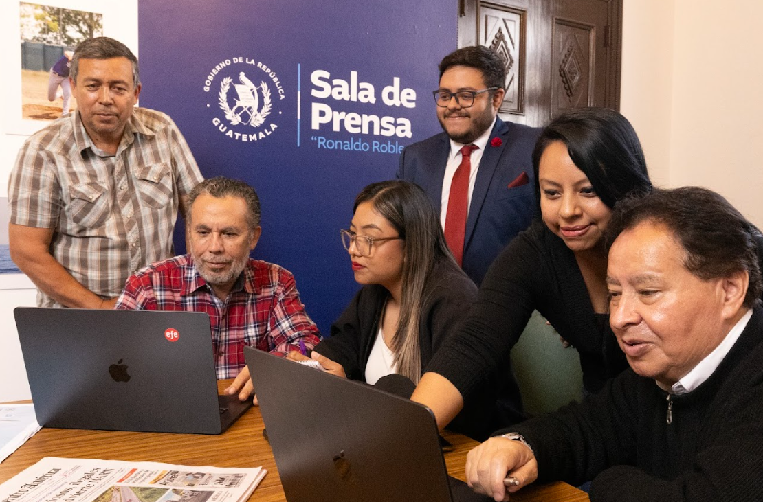 Seis personas reunidas alrededor de dos computadoras portátiles frente al rótulo de la Sala de Prensa Ronaldo Robles.