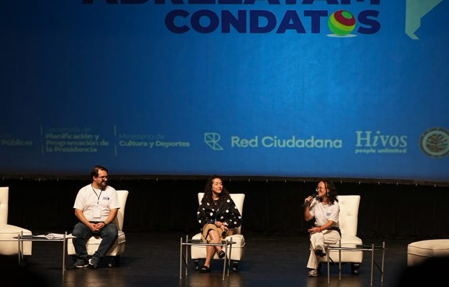 Tres panelistas conversan en un escenario frente a una pantalla con el nombre de Abrelatam/ConDatos.