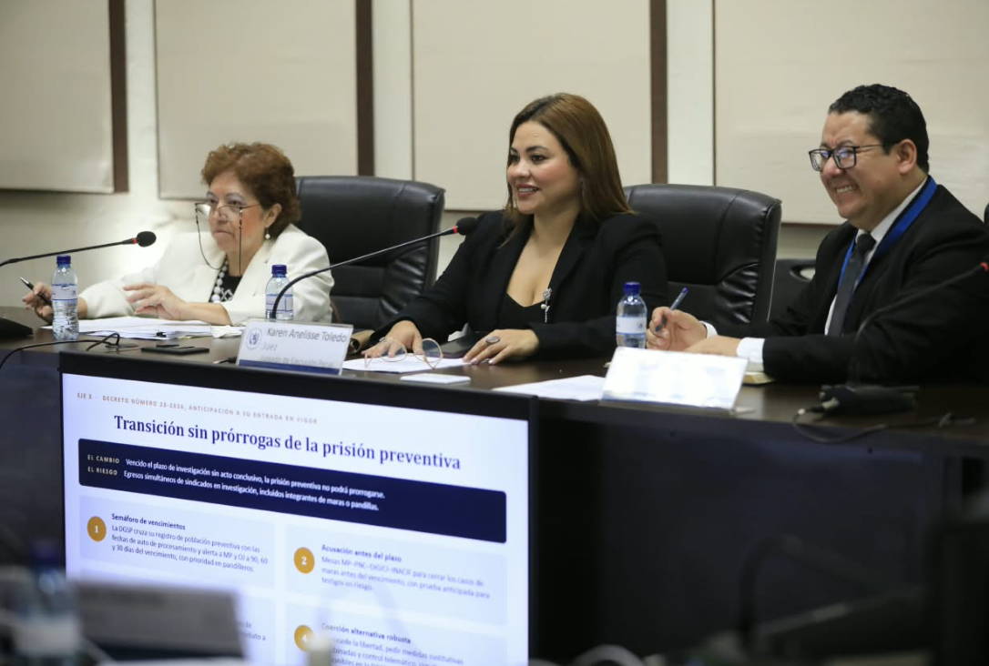 Tres participantes sentados frente a documentos y micrófonos, con una pantalla que muestra una presentación sobre prisión preventiva.