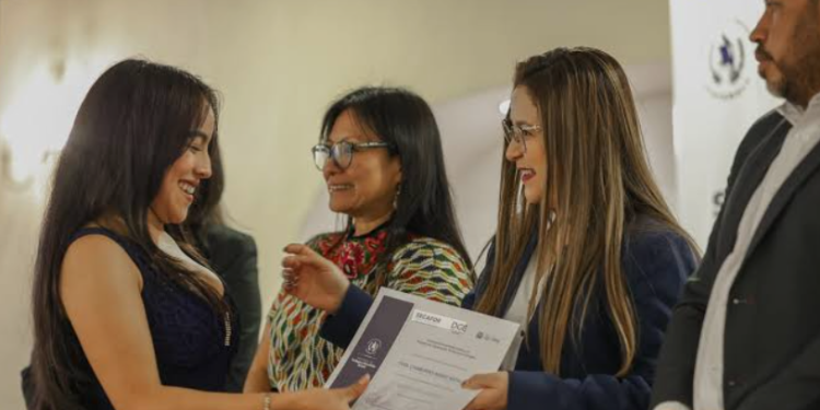 Una mujer recibe un diploma de manos de otra, acompañadas por dos personas durante una actividad institucional.