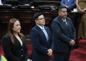 Tres personas con vestimenta formal permanecen de pie en el hemiciclo del Congreso de la República.