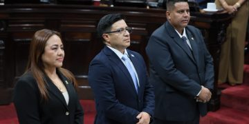 Tres personas con vestimenta formal permanecen de pie en el hemiciclo del Congreso de la República.