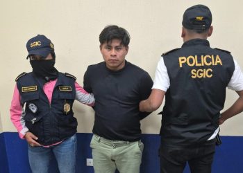 Hombre con camiseta negra y pantalón verde claro bajo custodia de dos agentes con chalecos de la PNC.