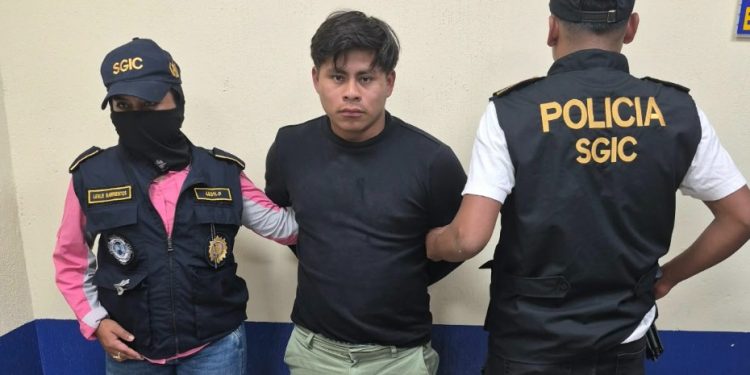 Hombre con camiseta negra y pantalón verde claro bajo custodia de dos agentes con chalecos de la PNC.