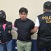 Hombre con camiseta negra y pantalón verde claro bajo custodia de dos agentes con chalecos de la PNC.