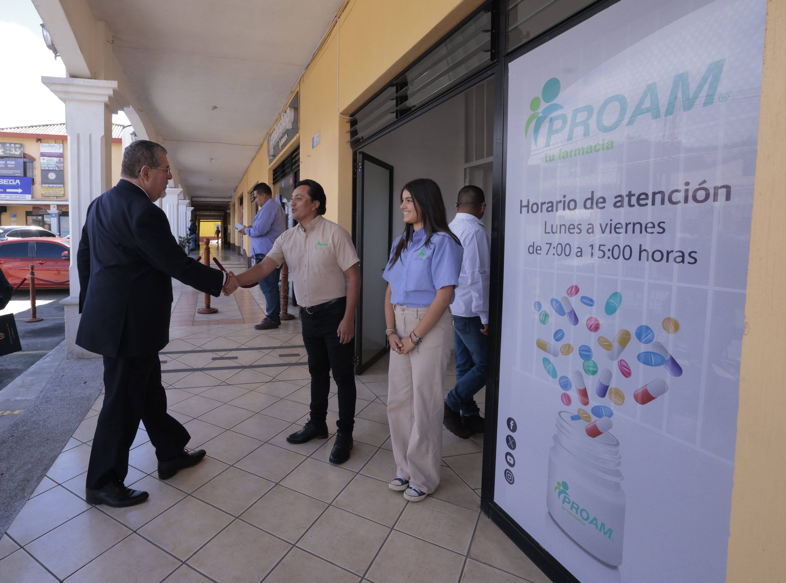 Bernardo Arévalo estrecha la mano de un trabajador frente a la farmacia, junto al rótulo con el horario de atención.