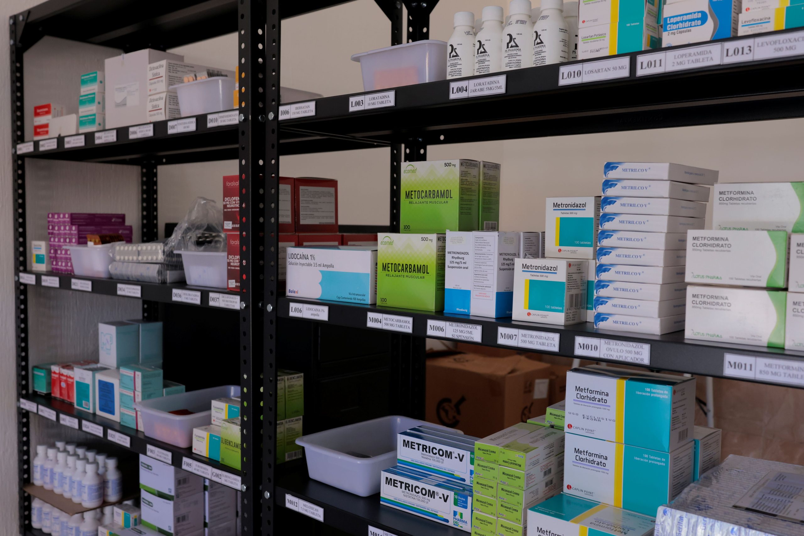 Cajas y frascos de medicamentos ordenados en estanterías metálicas con etiquetas de identificación.