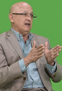 Hugo Maul, con anteojos y saco de cuadros, gesticula con ambas manos frente a un fondo verde.