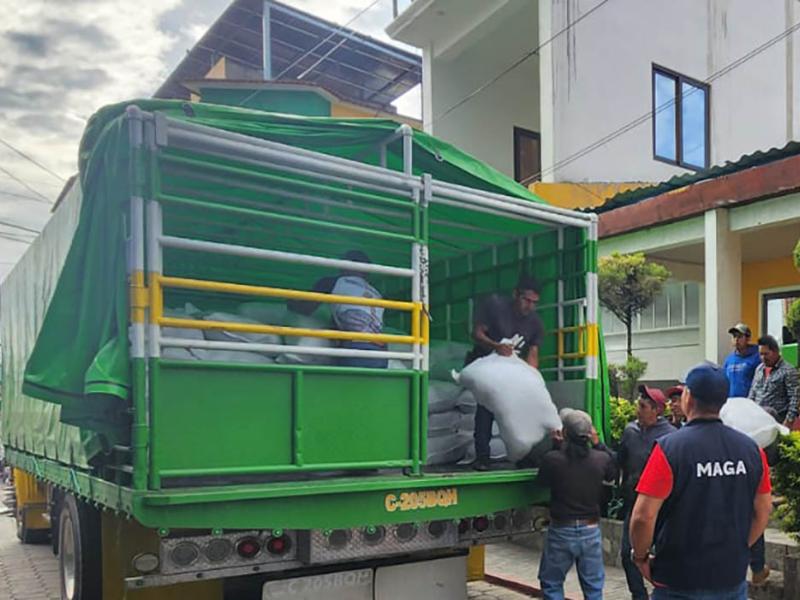 Varias personas descargan sacos de un camión verde, junto a un trabajador con chaleco del MAGA.