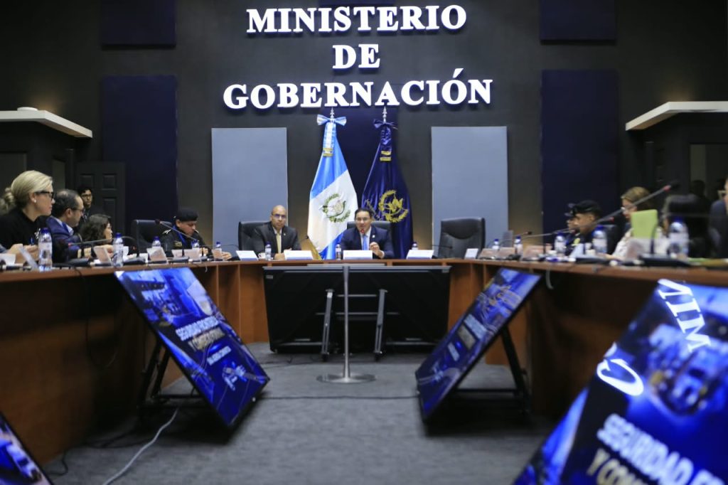 Participantes reunidos alrededor de una mesa en el Ministerio de Gobernación, con pantallas y banderas al frente.