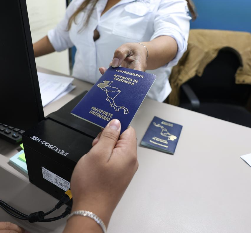 Dos personas sostienen un pasaporte guatemalteco sobre un escritorio con equipo de cómputo y otro pasaporte.