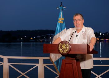 Bernardo Arévalo habla desde un atril con el escudo nacional, junto a la bandera de Guatemala y con un lago al fondo.
