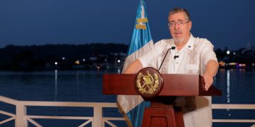 Bernardo Arévalo habla desde un atril con el escudo nacional, junto a la bandera de Guatemala y con un lago al fondo.