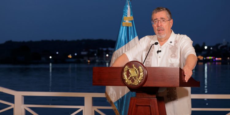 Bernardo Arévalo habla desde un atril con el escudo nacional, junto a la bandera de Guatemala y con un lago al fondo.