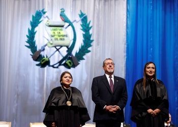 Bernardo Arévalo permanece de pie entre dos magistradas con toga, frente al escudo nacional y cortinas azules y blancas.