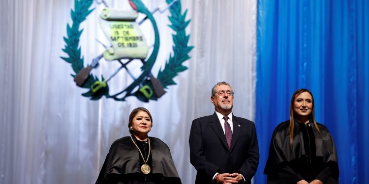 Bernardo Arévalo permanece de pie entre dos magistradas con toga, frente al escudo nacional y cortinas azules y blancas.