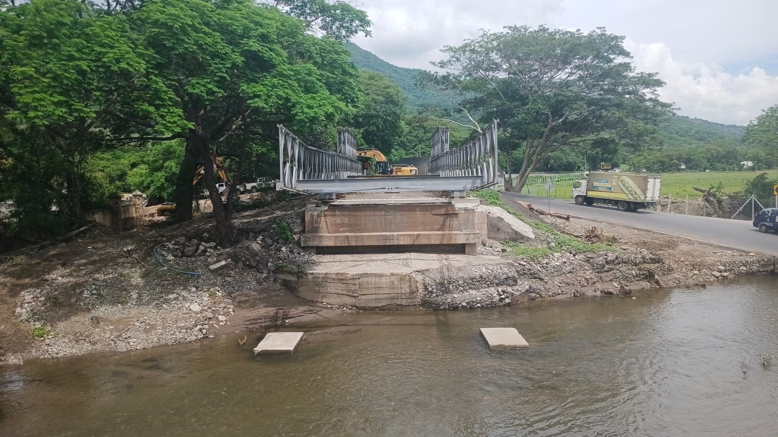 Estructura de un puente modular en montaje junto al cauce de un río, con maquinaria al fondo y vehículos en una vía lateral.