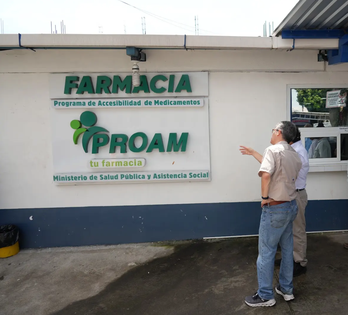 Joaquín Barnoya y un acompañante frente al rótulo de la farmacia del Programa de Accesibilidad de Medicamentos.