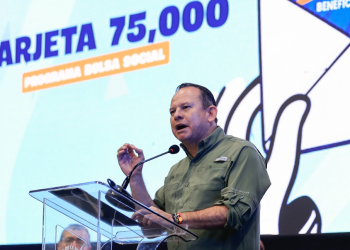 Abelardo Pinto habla ante un atril transparente, con una pantalla que anuncia la entrega de la tarjeta 75 mil del programa Bolsa Social.